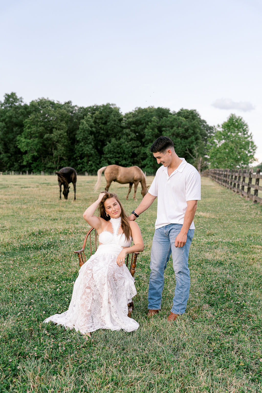 Bride in lace gown seated on wooden chair with horses grazing behind her, Bucks County farm